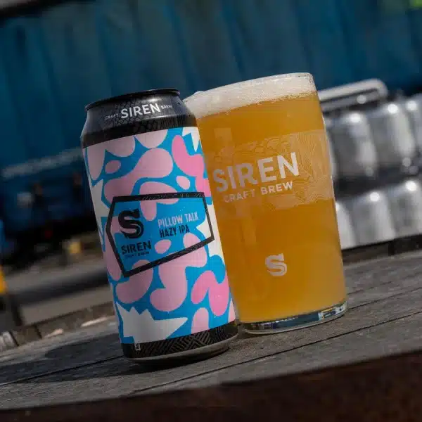 Two Siren craft beer items on a wooden table: a colorful Pillow Talk Hazy IPA can beside a glass of pale beer labeled SIREN Craft Brew, outdoors.